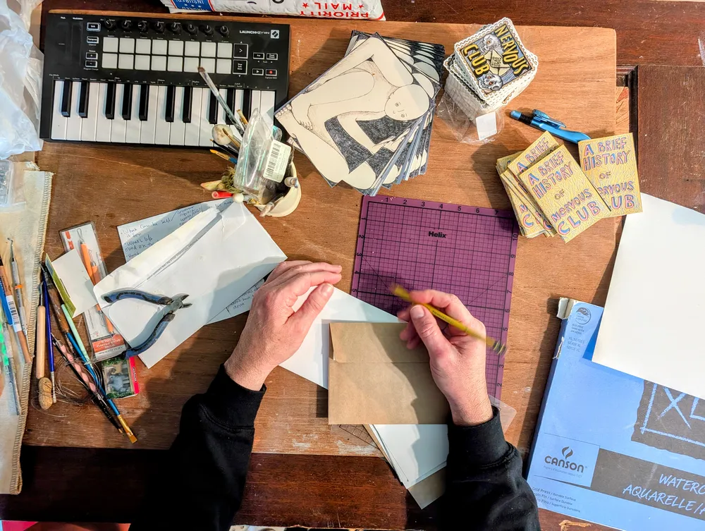 Top-down view of Ben's worktable — hands drawing on a cutting mat, surrounded by art supplies, envelopes, zines, and Nervous Club stickers